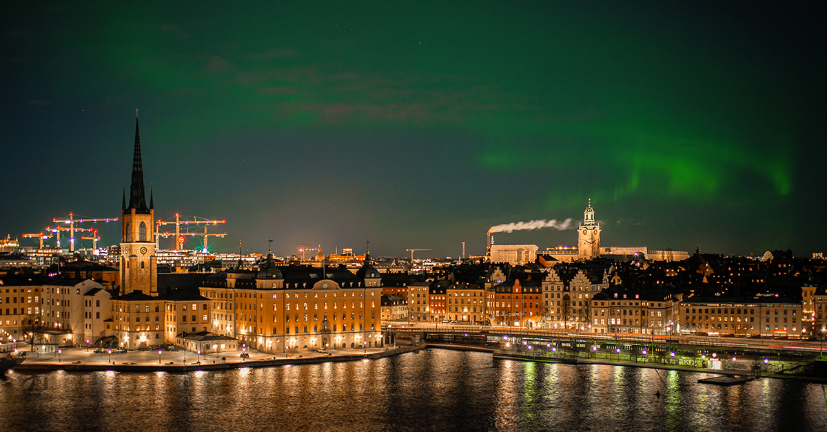 Night view of a city waterfront with lit buildings, a tall church spire, and green northern lights in the sky.
