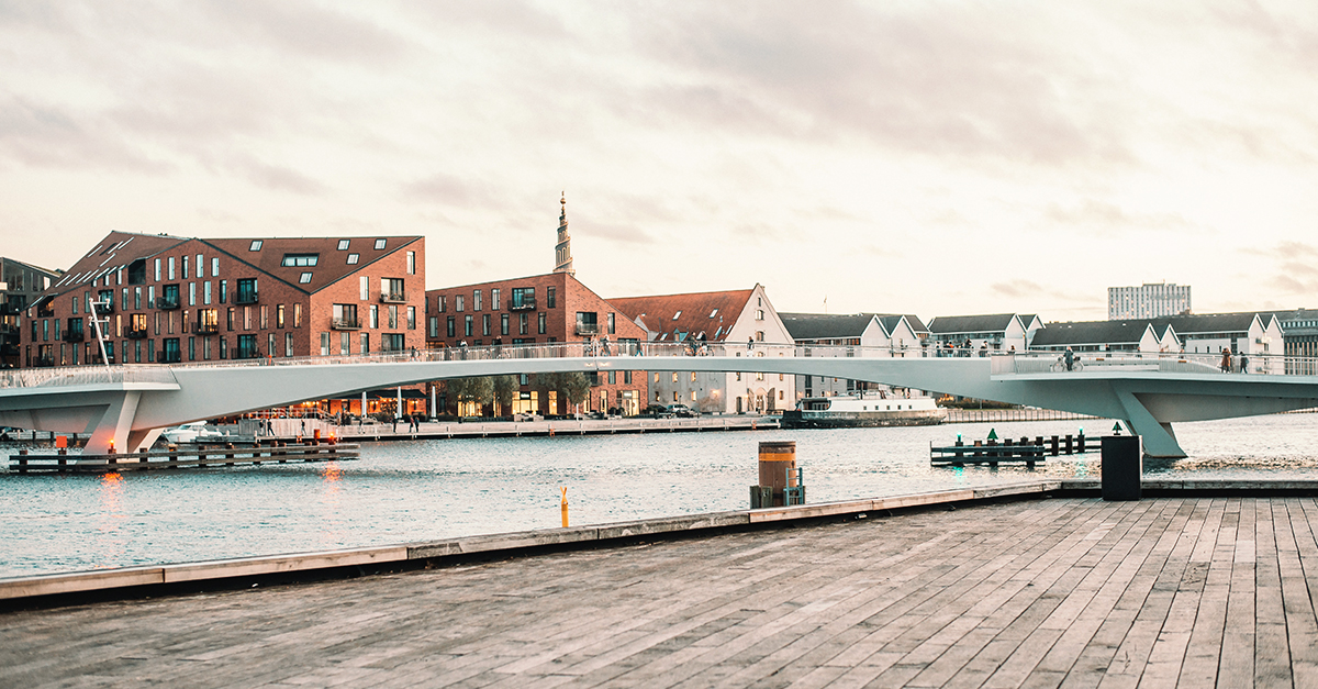 Modern pedestrian bridge over a canal with brick buildings and a spire in the background at sunset.