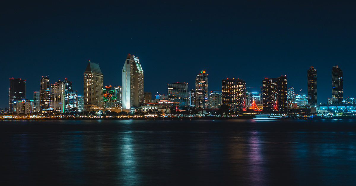 Night view of a city skyline with illuminated buildings reflecting on the water.