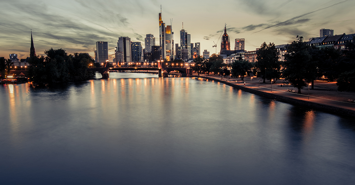 City skyline at dusk with illuminated buildings, a river, and a bridge reflecting city lights.