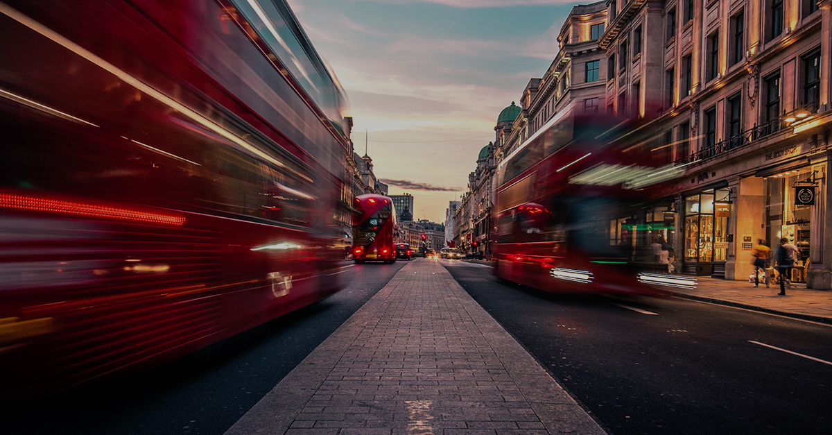 Blurred red double-decker buses move along a busy London street at dusk, flanked by historic buildings.