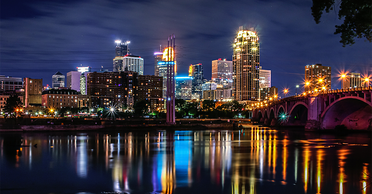 City skyline at night with illuminated buildings, a bridge, and colourful reflections on a calm river.