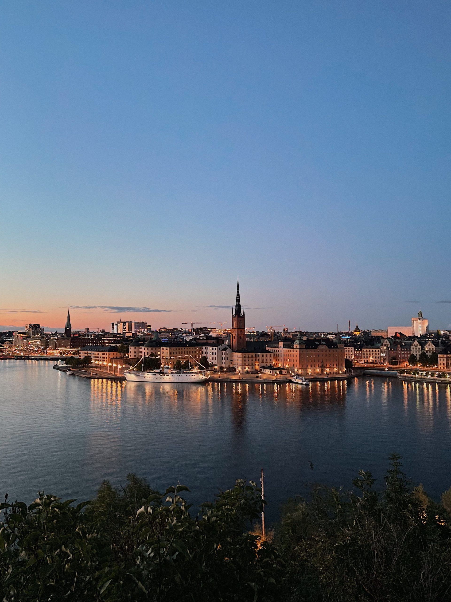City skyline at dusk with buildings and a church reflecting in calm water, lights shining along the waterfront.