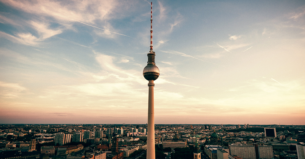Berlin TV Tower rises above city buildings under a partly cloudy sky at sunset.