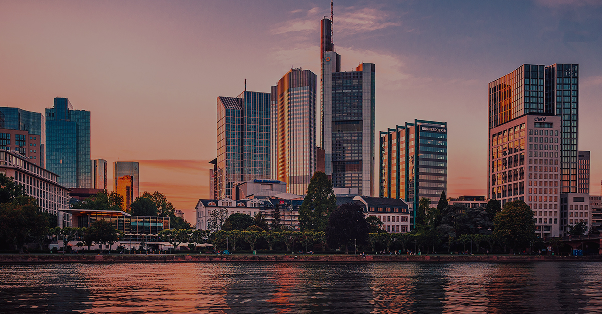 City skyline with modern skyscrapers at sunset, reflected on a calm river in the foreground.
