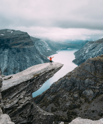 Person in a red jacket standing on a rocky cliff ledge above a fjord, surrounded by mountains and cloudy skies.