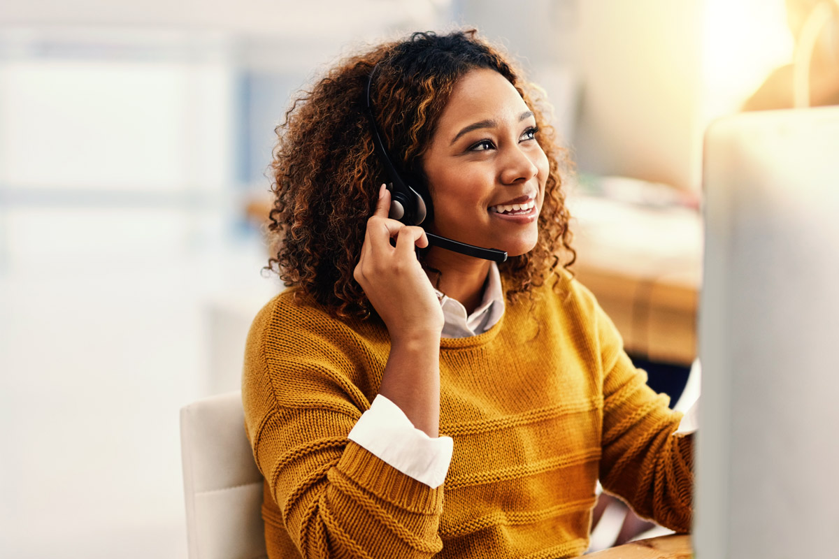 Woman in yellow jumper smiling, wearing a headset, sitting at a desk with a computer in an office setting.