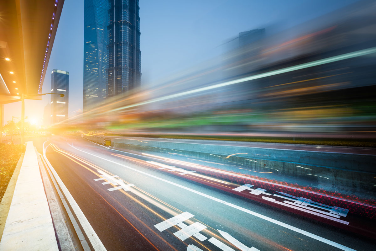 Blurred light trails from vehicles on a city road with tall skyscrapers at dusk.