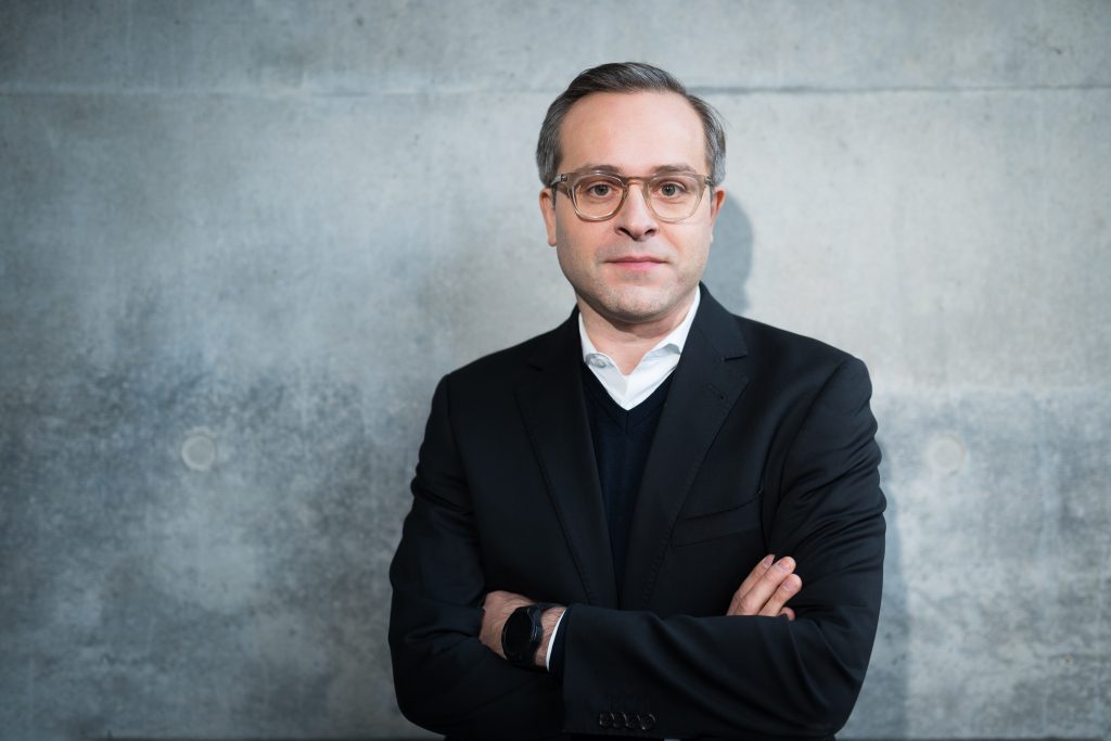 Man in a black suit with glasses standing with arms crossed against a plain grey wall.