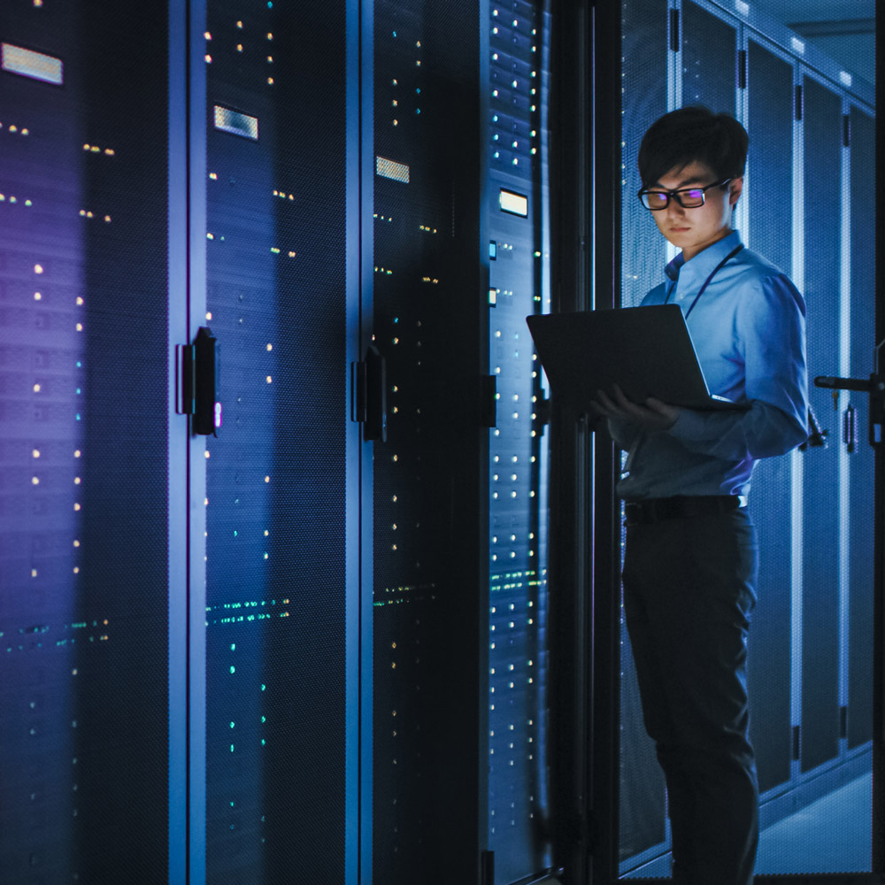 Person with a laptop standing in a server room with blue-lit server racks in the background.