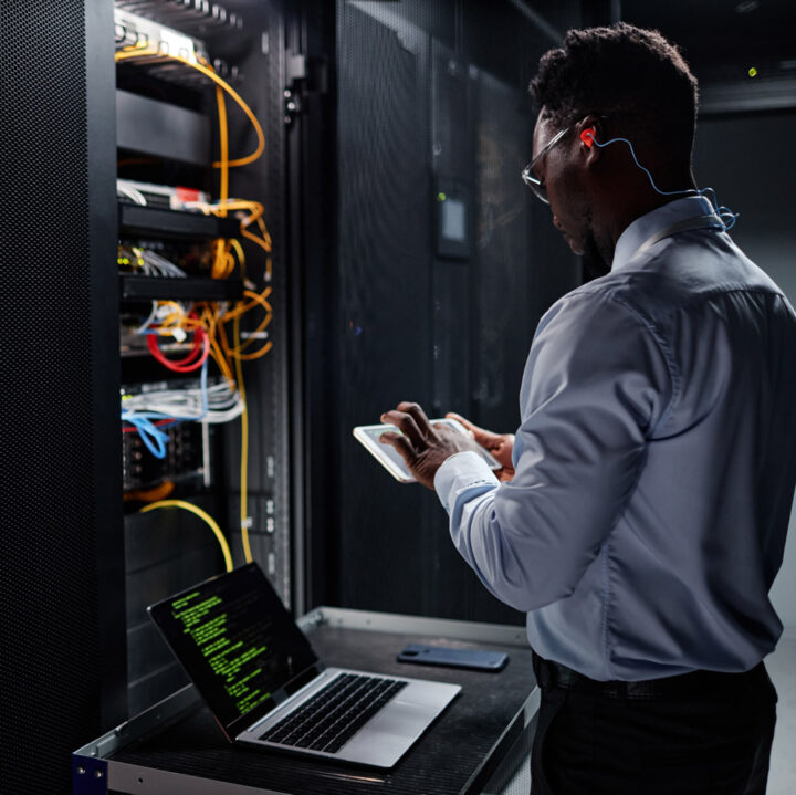 Man in server room working on tablet, laptop with code on screen, network cables and server racks visible.