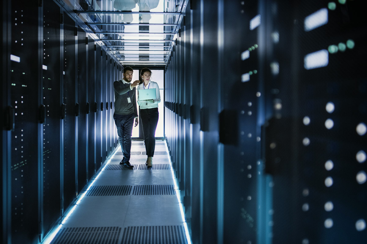 Two people walk in a server room corridor with racks on both sides, one holding a laptop, discussing work.