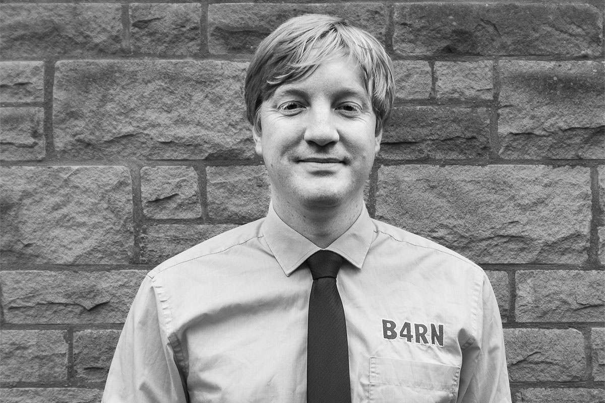 Man in shirt and tie with "B4RN" logo, standing in front of a brick wall, black and white photo.