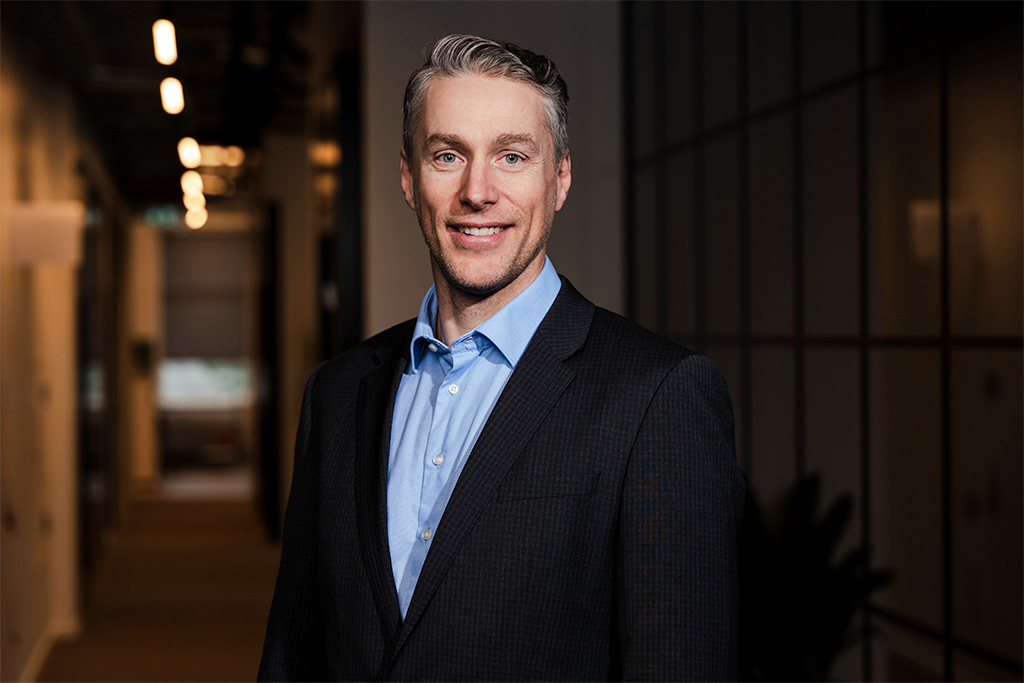 A man in a dark suit and blue shirt smiling, standing in a modern office corridor.