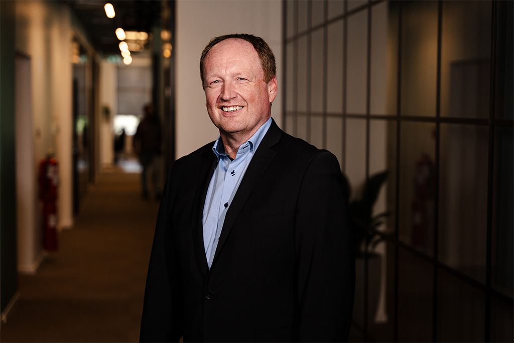 Smiling man in a dark suit jacket and blue shirt standing in a modern office hallway.
