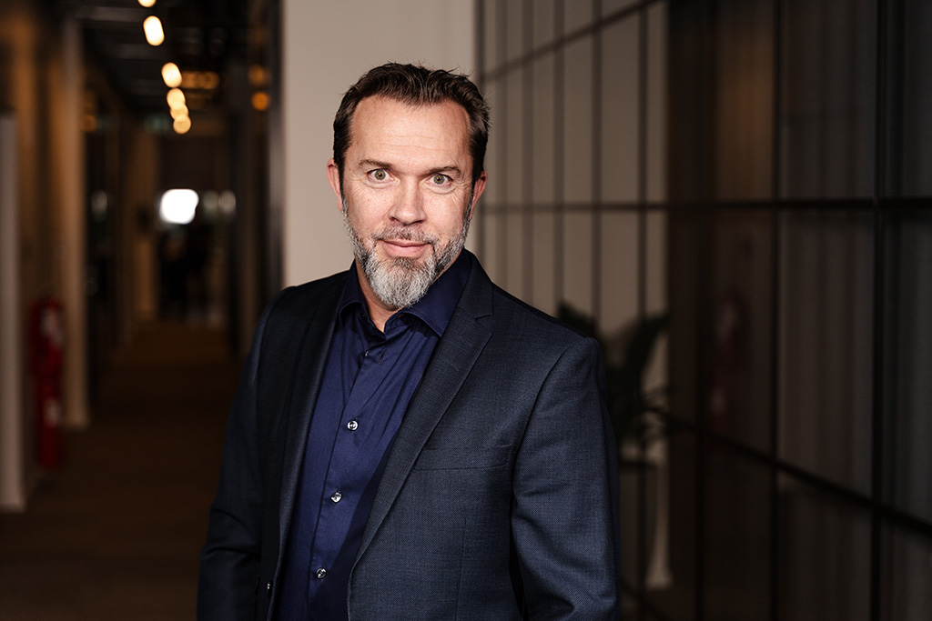 Man with short hair and beard wearing a dark suit, standing in a modern office hallway.