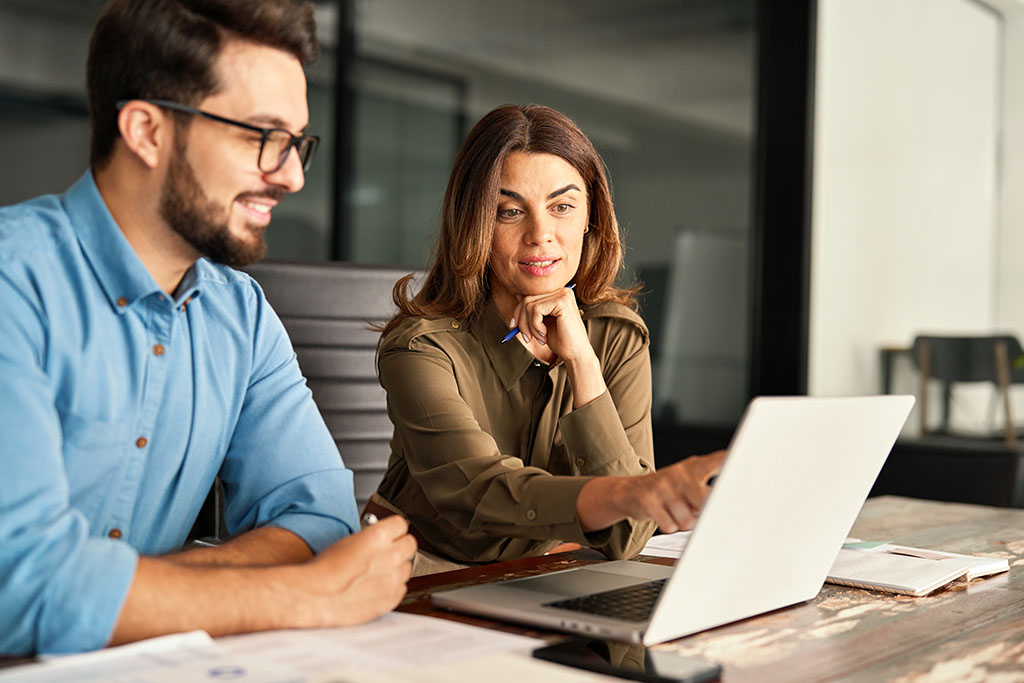 Two colleagues working together at a desk, looking at a laptop and discussing something.