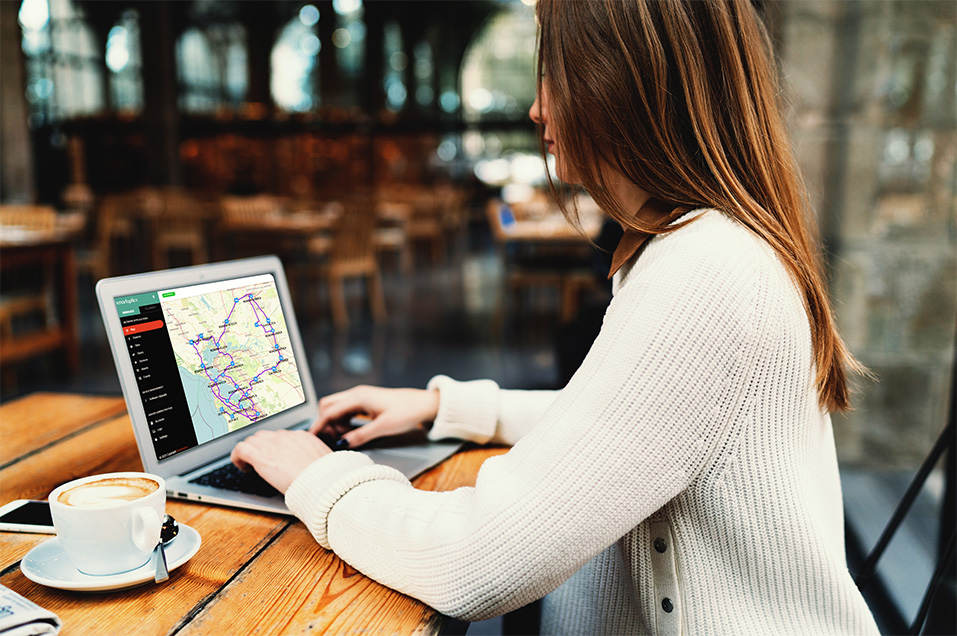 Woman using a laptop with a map on screen at a café table, coffee cup and phone beside her.