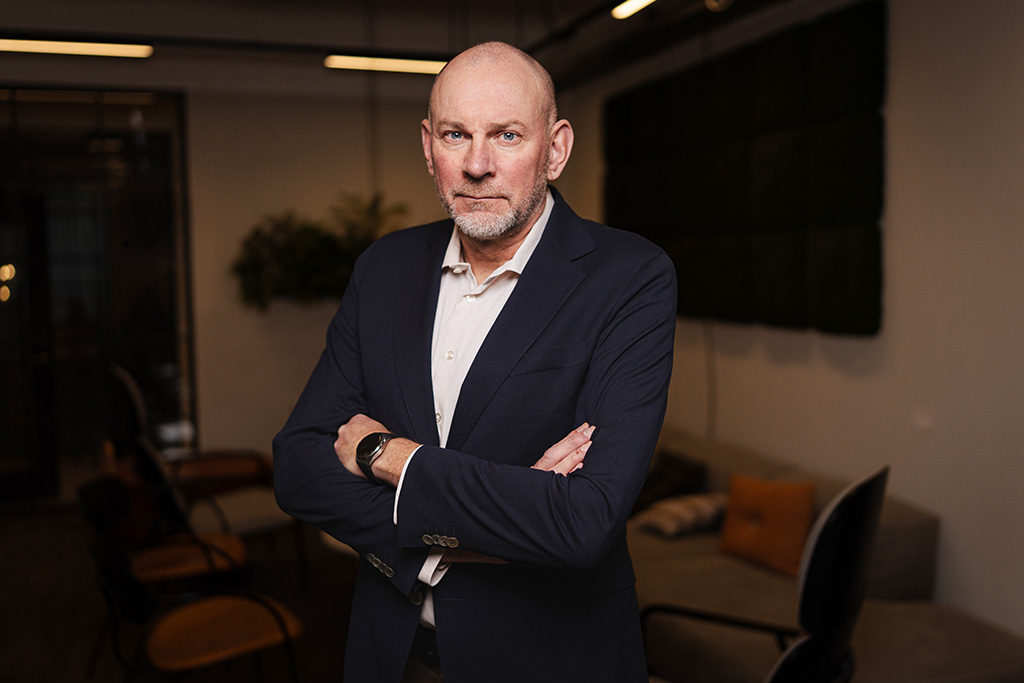Bald man in a suit with arms crossed, standing in a modern office with a sofa and chairs in the background.