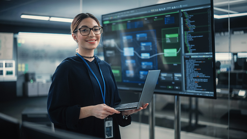 Woman with glasses holding a laptop, standing in front of large screen with code and data charts in office.